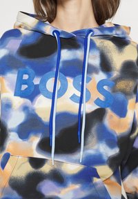 Felpa con cappuccio caratterizzata da un motivo tie-dye nei colori blu, arancione e nero, con un prominente logo "BOSS" blu e lacci a contrasto.
