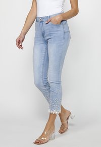 Light blue denim jeans with a fitted design, featuring floral lace embroidery at the hem. Includes front pockets and a button closure.