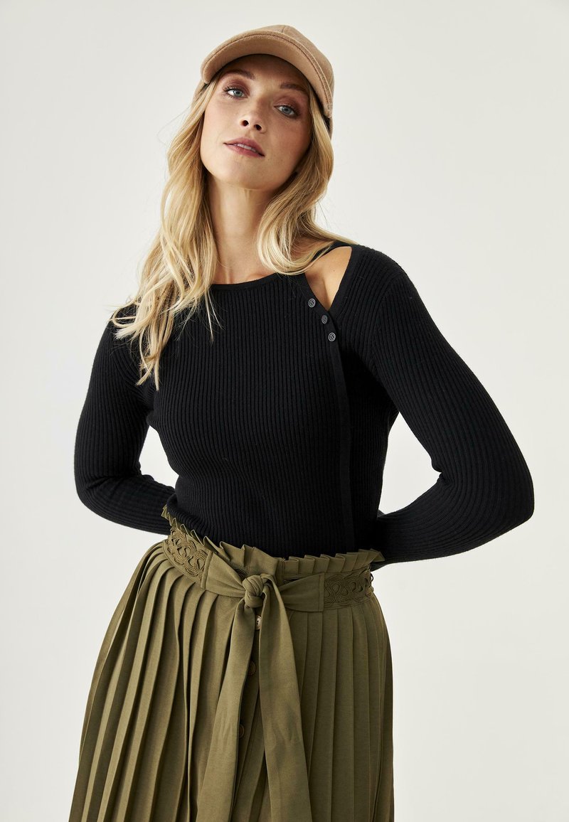 Suéter negro con canalé, con un detalle de corte en el hombro y botones. Combinado con una falda verde oliva con pliegues, que presenta una cinturilla decorativa y un lazo.