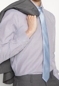 Chemise habillée avec un petit motif à carreaux violets, associée à une cravate bleu clair. Veste de costume grise drapée sur l'épaule, mettant en valeur un tissu texturé.