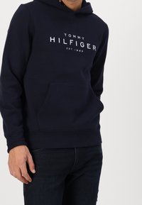 Uomo che indossa una felpa navy blue di Tommy Hilfiger con tasca frontale e jeans scuri, in piedi contro uno sfondo chiaro e uniforme.