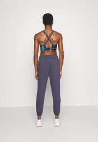 Sports-bh med blå og sort abstrakt mønster, der har krydsede stropper. Lilla sweatpants med elastiske manchetter og sidelommer. Hvide sneakers.