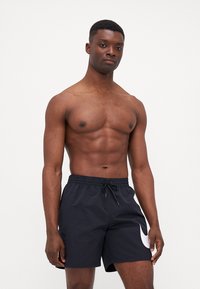 Pantaloni da nuoto neri con cintura elastica e cordino, caratterizzati da un grande logo Nike bianco sul lato sinistro. Texture liscia, design casual.
