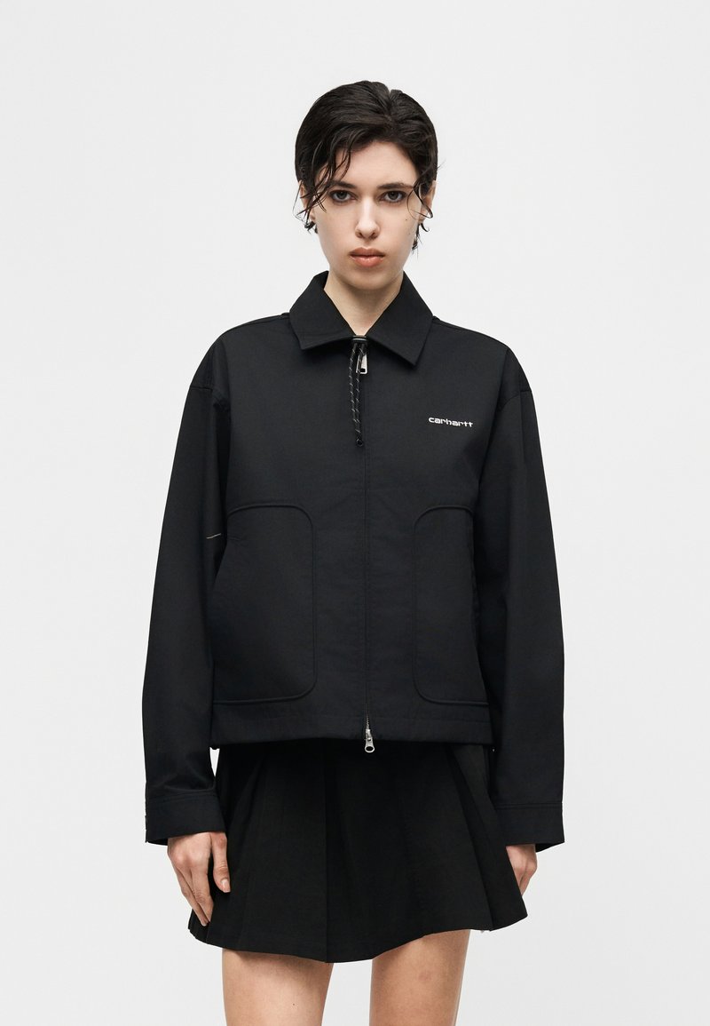 Persona con capelli corti e scuri che indossa una giacca nera con zip e logo "carhartt" e una gonna nera, in piedi contro uno sfondo chiaro e uniforme.