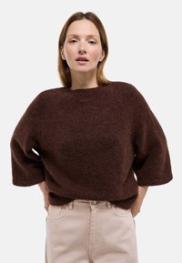 Femme aux cheveux bruns raides portant un pull ample en peluche de couleur brun foncé et un pantalon taille haute beige clair sur un fond uni.