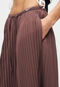 Καφέ αθλητικό παντελόνι με πιέτες, ελαστική μέση και κορδόνι. Διαθέτει πλαϊνές τσέπες και μικρό λογότυπο Nike στην μέση.