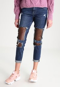 Oslabené džínové kalhoty s hnědými umělými koženými nášivkami na kolenou, ohrnutými lemy a modrým odbarvením. Doplňují je růžové tenisky.