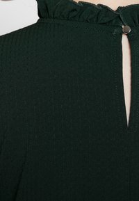 Tmavě zelená látka šatů s ribkovanou strukturou, s volánkovým límcem a zapínáním na klíčovou dírku vzadu s malým kovovým knoflíkem.