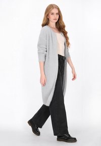 Hellgrauer gestrickter Cardigan mit langem, offenem Design, gerippter Textur und umgeschlagenen Ärmeln, kombiniert mit schwarzen, weiten Hosen und schwarzen Schuhen.