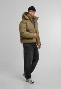 Puffjakke i olivengrønn, med høy krage og hette, glidelås-lukking, kombinert med mørke jeans og sølvfargede sneakers.