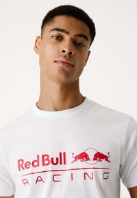 Witte katoenen t-shirt met daarop de tekst "Red Bull Racing" in het rood, vergezeld van twee rode stierafbeeldingen boven een cirkelvormig ontwerp. Korte mouwen, ronde hals.