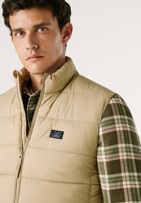 Gilet imbottito beige con colletto alto, chiusura a zip e patch del marchio. Indossato sopra una camicia di flanella a quadri verde con dettagli rossi.