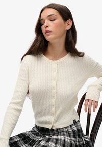 Kremowy kardigan z prążkowanej dzianiny wykonany z miękkiego materiału, z okrągłym dekoltem, sześcioma guzikami i długimi rękawami. W komplecie ze spódnicą w kratę.