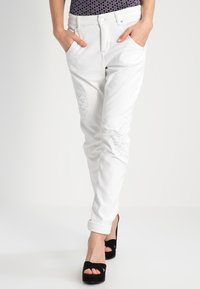 Białe dżinsy z przetarciami, w slim fit, z podwiniętymi nogawkami i kieszeniami z przodu, połączone z czarnymi sandałami na obcasie.