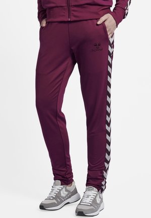 Persoon draagt bordeauxrode trainingsbroek met witte chevronstrepen aan de zijkanten en grijs-witte sneakers, staand met handen in de zakken.