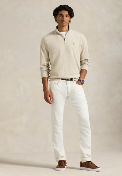 Beige genser med glidelås i halsen, hvite jeans, brune sko; glatt stoff, avslappet passform, subtil logo på genseren, kontrastsøm på jeansene.