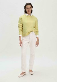 Pull en laine jaune flou avec un col rond, chemise blanche à col en dessous, associé à un jean blanc taille haute et des ballerines beige clair.
