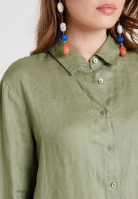 Jasnozielona lniana koszula z kołnierzykiem i zapięciem na guziki. Charakteryzuje się subtelną fakturą. Akcesoria to kolorowe kolczyki z koralików.