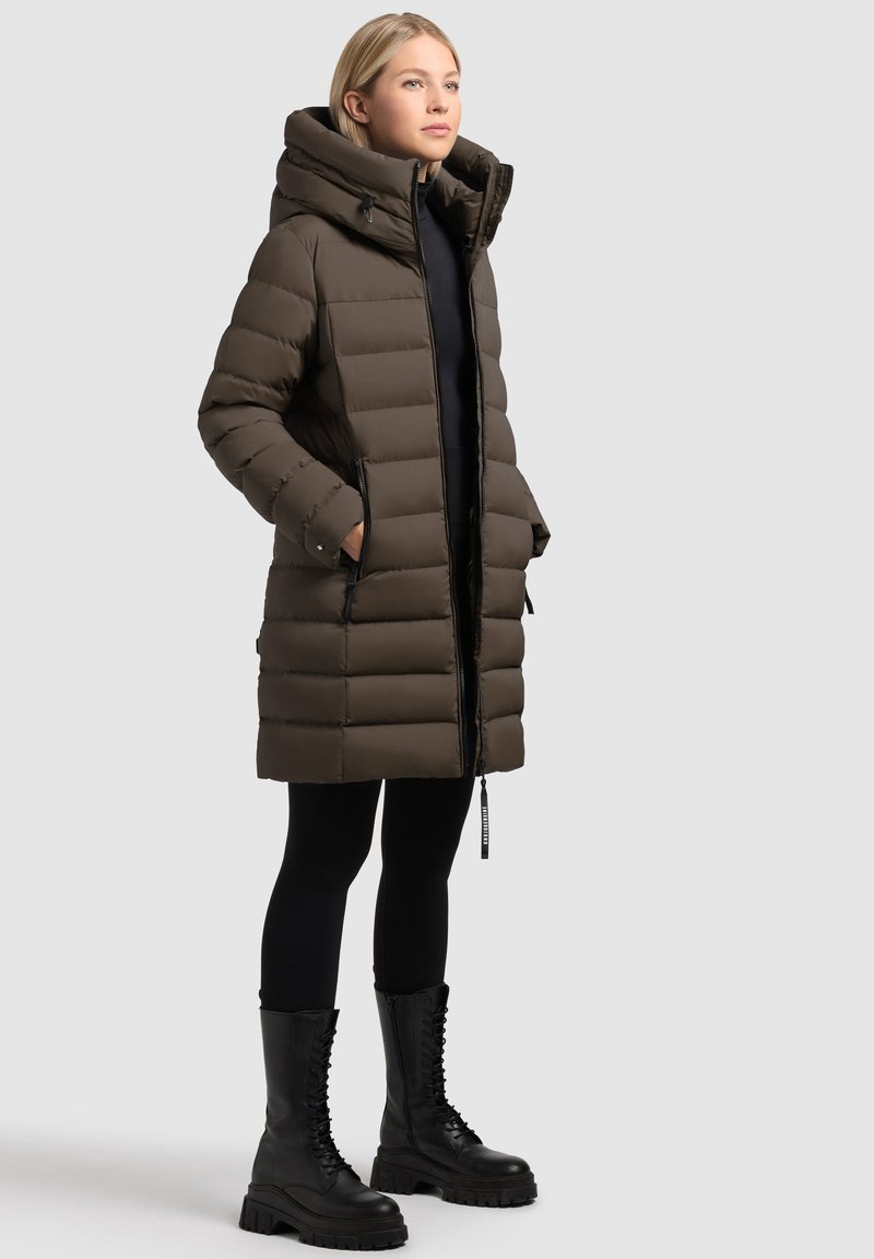Vadderad brun jacka med hög krage och huva, med dragkedjefickor och knälång längd. Bärs med svarta leggings och snörkängor.
