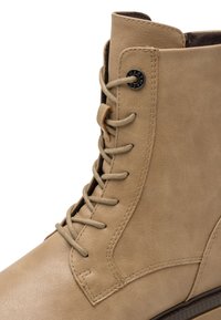 Bottine en cuir beige à lacets, à bout arrondi et œillets noirs. Présente une finition texturée et de légères coutures décoratives.