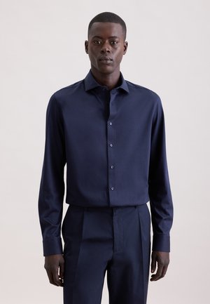 Mężczyzna ubrany w granatową, długorękawową koszulę na guziki i dopasowane granatowe spodnie, stojący na neutralnym jasnym tle.