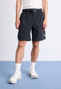 Person wearing black cargo shorts with belt, white crew socks, and white sneakers standing on blue floor against plain wall.