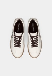 Λευκά δερμάτινα αθλητικά παπούτσια με καφέ κορδόνια, με μινιμαλιστικό σχεδιασμό και ραβδωτή σόλα. Το λογότυπο είναι ορατό στη γλώσσα.