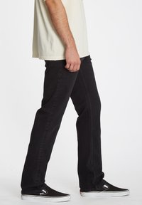 Zwarte denim jeans met een recht model, voorzien van subtiele stiksels, gecombineerd met zwarte instapschoenen, zijwaarts weergegeven.