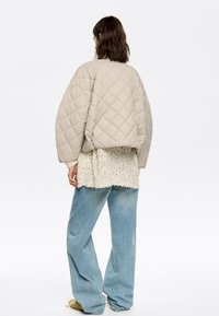 Beige quilted jacket with a cropped silhouette, paired with a textured white top and loose light blue jeans. Soft, smooth material.