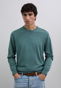 Grön långärmad tröja med rund halsringning, gjord av mjukt tyg, kombinerad med ljusblå denimjeans och ett brunt flätat skärp.