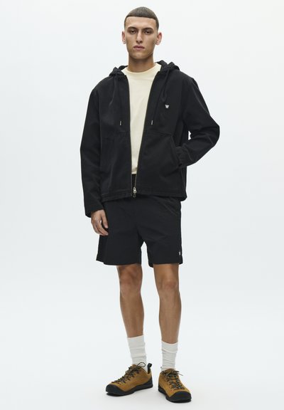 Veste noire à capuche, chemise beige, short noir et chaussures de randonnée marron. Design minimaliste avec des poches et un petit logo sur la veste.