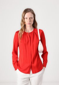 Rød langermet bluse med knappet hals, glatt stoff, plisserte detaljer, sammen med en liten rosa veske hengende fra skulderen.