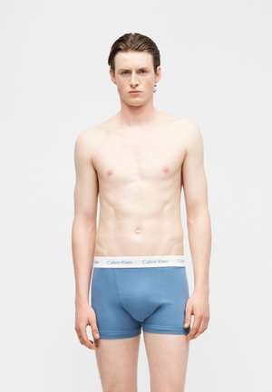 Młody mężczyzna o krótkich brązowych włosach stoi na białym tle, ubrany w niebieskie bokserki Calvin Klein.