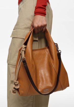 Mano sosteniendo un bolso de cuero color canela con detalles de cuerda beige y correa marrón oscuro, combinado con pantalones cargo beige y una camiseta de manga larga roja.