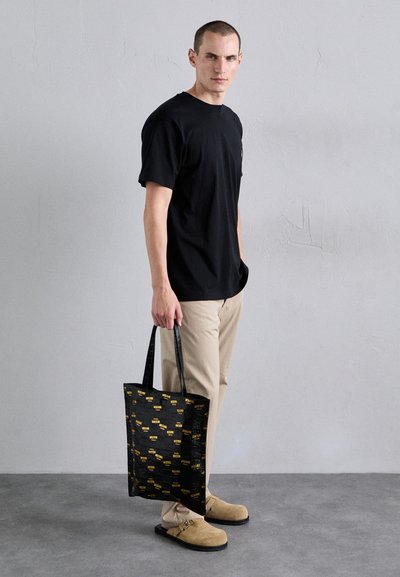 Geantă tote neagră cu branding galben, textură lucioasă și mânere negre. Modelul poartă un tricou negru și pantaloni bej, cu încălțăminte de culoare deschisă.