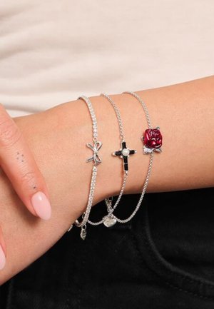 Braccialetti d'argento con charm: un fiocco rosa, una croce nera con una perla e una rosa rossa, su uno sfondo di pelle testurizzata.