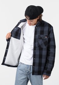 Black and navy plaid shirt-jacket, featuring a fuzzy white lining, button-up front, and chest pocket. Soft texture with a relaxed fit.