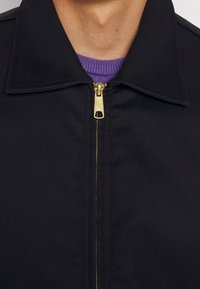 Svart zip-up jacka med platt krage; har en guldfärgad dragkedja och en synlig lila ribbad trim vid nacken. Slät, strukturerad tyg.