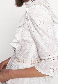 Valkoinen, broderad puuvillamekko, jossa on kerroksittain laskoksia, kukkakuosisia ja pitsikoristeluja pääntiellä ja hihoissa. Koristeelliset leikkaukset näkyvät.