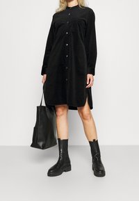 Czarna sukienka koszulowa z korduroy z przodem na guziki, długimi rękawami, szczelinami po bokach i kieszenią na piersi. Noszona z czarnymi botkami na kostkę i torebką. 