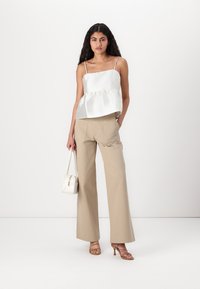 Hvit sateng peplum-top med tynne stropper, kombinert med beige vidde bukser. Modell som holder en liten hvit håndveske, ikledd brune høye sandaler.