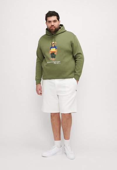 Zelená mikina s kapucňou a grafickým dizajnom, biely potlačený logo, v kombinácii s bielymi šortkami a bielymi teniskami. Jednoduchý, neformálny športový outfit.
