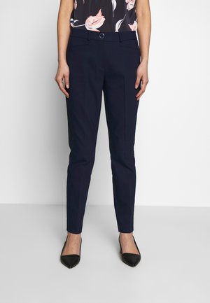 Kvinde iført slim fit marineblå bukser, blomstret ærmeløs top og sorte spidse sko, der står mod en hvid baggrund.