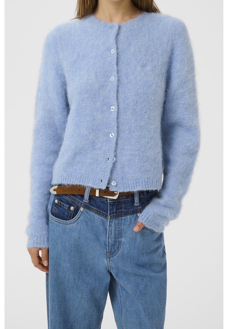 Femme portant un cardigan bleu clair, duveteux et boutonné, à manches longues, rentré dans un jean bleu taille haute avec une ceinture marron.