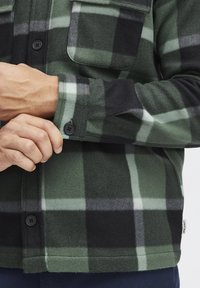 Camisa de franela a cuadros verde y negra con dos bolsillos en el pecho y puños de botón. Textura suave con patrones contrastantes en blanco y gris.