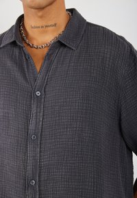 Camicia button-up grigia con texture e motivo a scacchi, dotata di colletto e bottoni blu. Include una collana in argento.