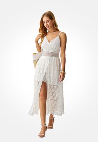 White lace maxi dress with a deep V-neck, adjustable straps, and patterned texture. Features a side slit and a crochet waist detail.