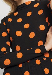 Femme portant un haut noir à manches longues côtelé avec de grands pois orange, croisant le bras à la taille.