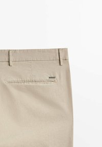 Beżowe spodnie widok z tyłu z szlufkami na pasek i jedną kieszenią wpuszczaną, z małą prostokątną metką marki umieszczoną nad kieszenią.