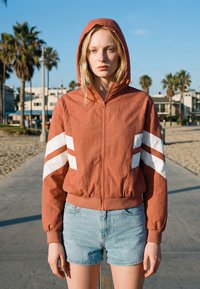 Jonge vrouw staat op een zonnig pad langs het strand, gekleed in een roestkleurige jas met capuchon met witte strepen en lichtblauwe spijkershorts.
